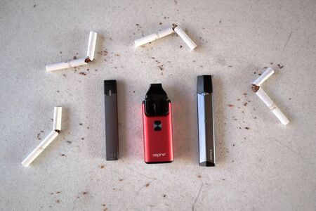 Different vape devices electronic cigarettes juul, aspire breeze, smok infinix, as smoking alternatives with broken marlboro gold cigarettes and scattered tobacco on white textured backgroundのeditorial素材