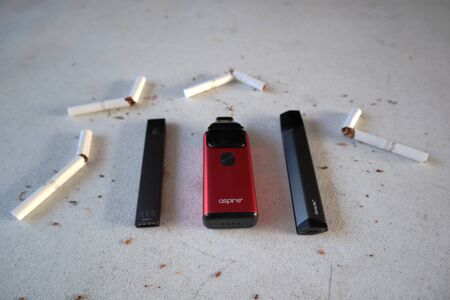Different vape devices electronic cigarettes juul, aspire breeze, smok infinix, as smoking alternatives with broken marlboro gold cigarettes and scattered tobacco on white textured backgroundのeditorial素材