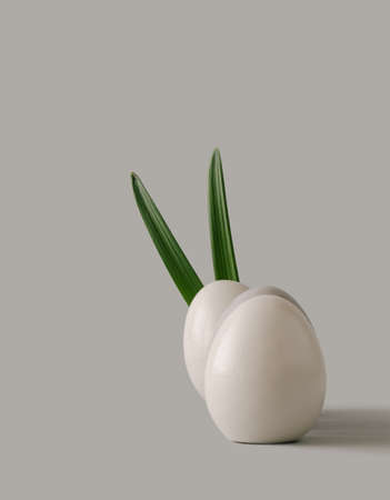 Easter white eggs with bunny ears made of natural green leaves on bright grey background. Easter minimal concept.の写真素材