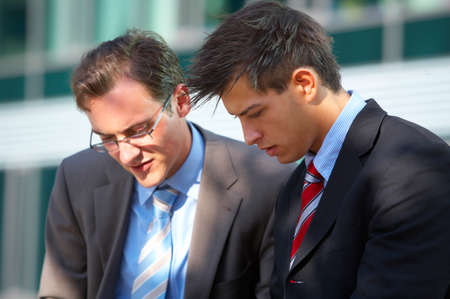Two young business men are working outside while sitting on bankの写真素材