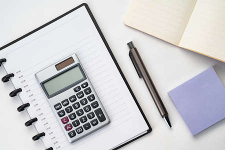 Top view of white table with notebook, calculator, pen and sticky note
