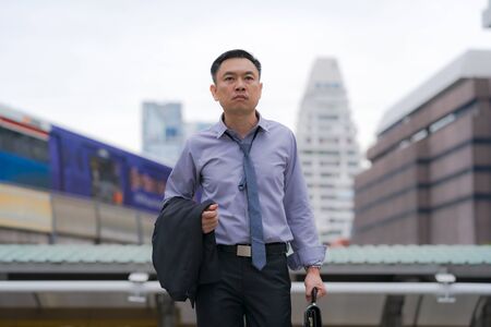 Asian Businessman walking and holding briefcase with business office buildings in the city backgroundの写真素材