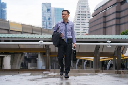 Asian Businessman walking and holding briefcase with business office buildings in the city backgroundの写真素材