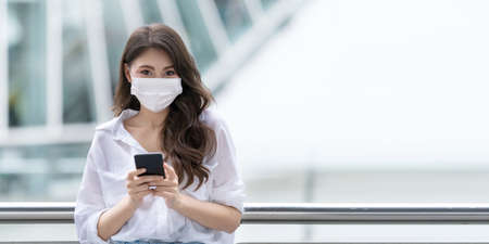 Portrait of young woman wearing medical face mask protective using a phone walks in a cityの写真素材