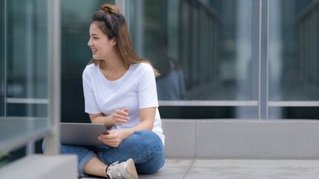 Woman in white t-shirt and blue jeans using laptop sitting on a floor outside in the city street background, Summer dayの写真素材