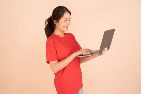 A beautiful Asian woman in a red shirt is holding a laptop computer on a cream background She is having fun surfing the web.の写真素材