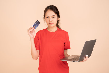A young woman wearing a red shirt holds a credit card and uses a laptop to pay online,online shopping, ecommerce, banking, internet cafes using credit card to pay.の写真素材