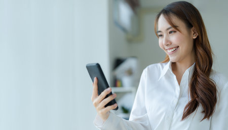 Happy young asian business woman wearing suit holding mobile phone standing in her workstation officeの写真素材