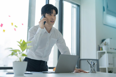 Asian businessman wearing suit holding mobile phone standing in workstation officeの写真素材