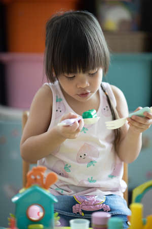 A three-year-old Asian girl with long black hair and a braid. which is an innocent and cute age, she is playing Kitchen-related toys that include cups, plates, bowls, fixing spoons and condiment sets. and was scooping up the food she had madeの写真素材