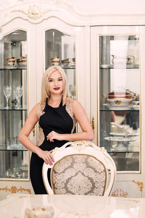 Stylish elegant blonde woman in beauty rich interior, wearing black dress. Luxurious intranier in white toneの写真素材