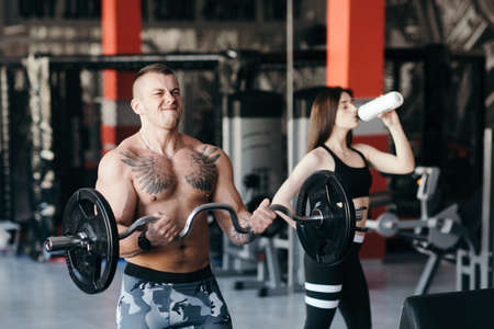 Strong man - bodybuilder with dumbbells in a gym, exercising with a barbell and on the background girl drinks waterの写真素材