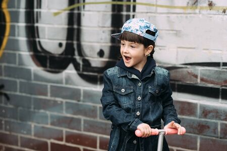 Cute little girl in jeans cardigan and on scooter on the background the urban colorful wall. Springの写真素材