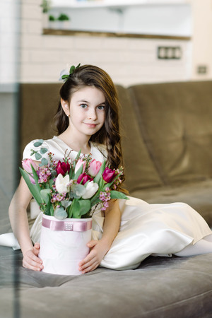 A cute girl sits on a sofa and holds flowers in her hands. Feast dayの写真素材