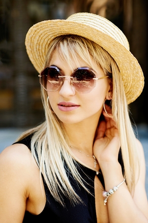 A beautiful blonde young girl in a black dress and straw hat sitting on the background of the door of the store and the granite wall.の写真素材