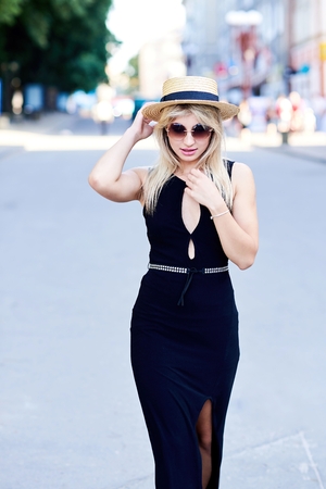 A beautiful blonde young girl in a black dress and straw hat walk in the summer sunny city.の写真素材