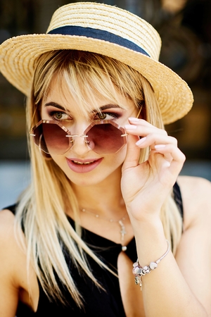 A beautiful blonde young girl in a black dress and straw hat sitting on the background of the door of the store and the granite wall.の写真素材