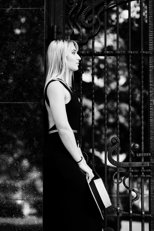 A beautiful blonde young girl in a black dress and straw hat poses on the background of the door of the store and the granite wall.の写真素材