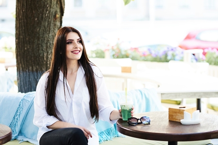 Cute young brunette girl with beautiful professional make-up sitting in a street cafe on a beautiful sunny dayの写真素材