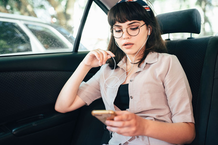 Beautiful young pretty girl is using a smart phone and smiling while sitting on back seat in the car.の写真素材