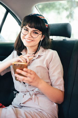 Beautiful young pretty girl is using a smart phone and smiling while sitting on back seat in the car.の写真素材