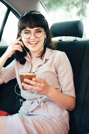 Beautiful young pretty girl is using a smart phone and smiling while sitting on back seat in the car.の写真素材