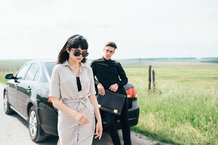 Couple of young girl and boy holding talks near black car on background of blue sky and field.の写真素材