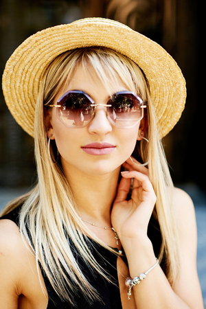A beautiful blonde young girl in a black dress and straw hat sitting on the background of the door of the store and the granite wall.の写真素材