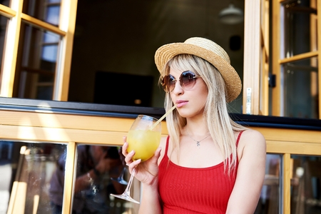 Young pretty sympathetic blonde sitting in a cafe near an open window with a cocktail in her arms and in a straw hat.の写真素材