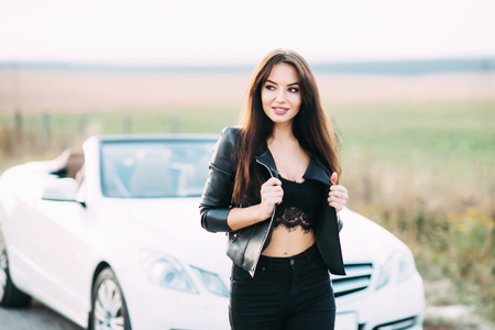 A beautiful woman dressed in trousers and a leather jacket waiting for a white cabriolet car.の写真素材