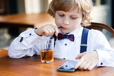 A cute white-haired boy, school years dressed in school uniform, spends his time at the summer cafe on the street. The guy drinks juice and works with his smartphoneの写真素材