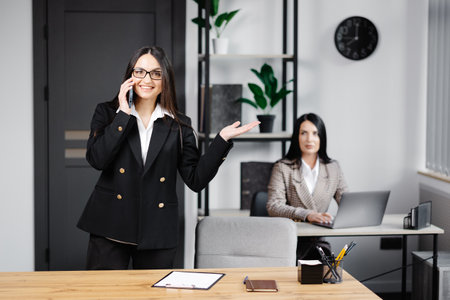 Attractive young business woman wearing jacket talking on mobile phone while sitting on a desk and using notebook near her colleague at work in office.の写真素材