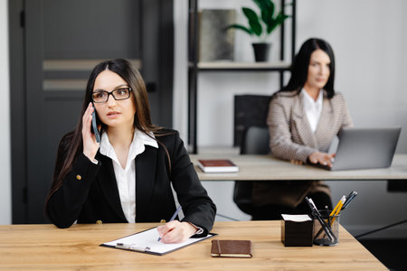 Attractive young business woman wearing jacket talking on mobile phone while sitting on a desk and using notebook near her colleague at work in office.の写真素材