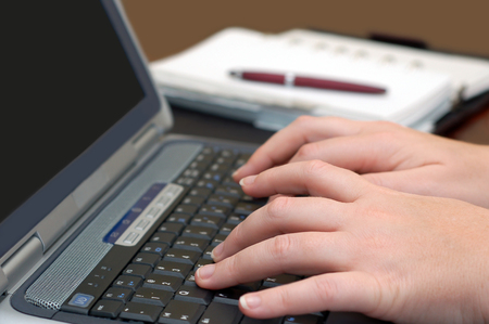 Notebook computer with hands typing and a day planner and pen in the background.の写真素材