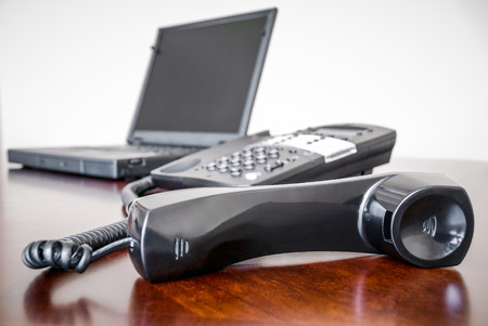 Picture of a black business telephone with its receiver off the hook and a laptop computer on a brown office deskの写真素材