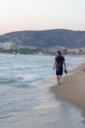 The young man is walking on the beach and has shoes in his hand.の写真素材