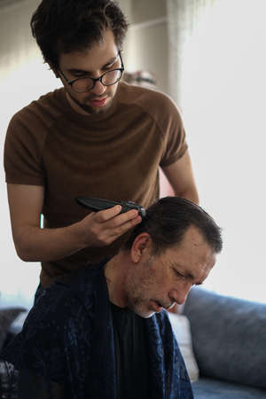 The young man is cutting father's hair with hair clipper during coronavirus quarantine isolation at home .Home haircut concept.の写真素材