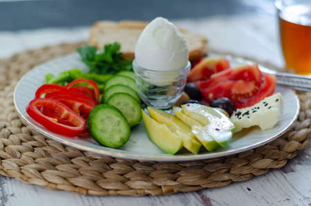 Healthy keto diet breakfast: boiled egg, avocado, cheese, cucumber, pepper, tomato, parsley, whole grain bread, and teaの写真素材