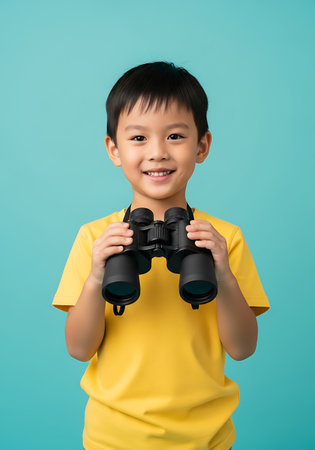 A young Asian boy wearing a bright yellow t-shirt smiles at the camera while holding a pair of black binoculars. His hands are wrapped around the binoculars, which hang from a black strap around his neck. The background is a solid, vibrant turquoise blue. The boy's expression is one of curiosity and excitement, suggesting a sense of adventure or discovery.の素材