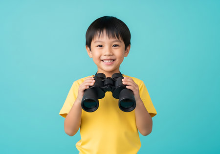 A young boy with black hair and a bright smile wears a yellow t-shirt and holds a pair of black binoculars in front of a solid blue background. His hands are clasped around the binoculars, and he appears to be looking forward with a sense of curiosity and excitement. The lighting is even, highlighting the subject against the plain backdrop.の素材