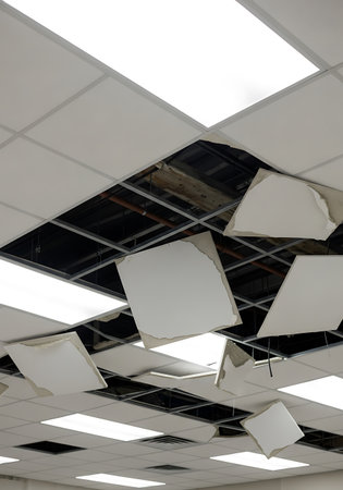 A suspended ceiling in an interior space shows significant damage. Several white ceiling panels are broken and hanging loosely from the grey metal grid structure. The exposed area reveals the dark ceiling cavity with visible pipes and structural elements. Some intact ceiling panels and illuminated light fixtures are also visible.の素材