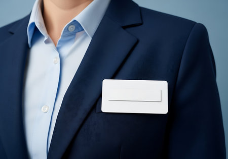 A close-up shot shows a person wearing a dark blue suit jacket over a light blue collared shirt. A white rectangular name tag with a blank insert is attached to the lapel of the suit jacket. The background is a solid light blue.の素材