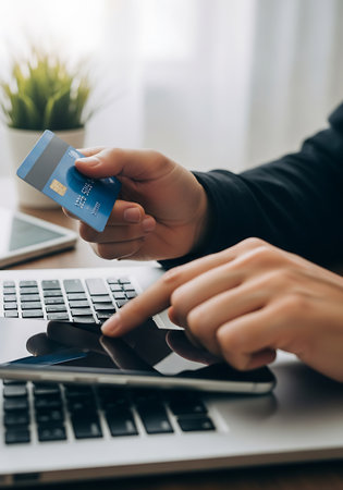 A person's hands are shown interacting with a laptop and a credit card. One hand holds a blue credit card with visible numbers and a chip. The other hand is positioned over the laptop's keyboard, with a finger touching a tablet or screen that rests on the keyboard. The background is blurred, showing a green potted plant and a bright window.の素材