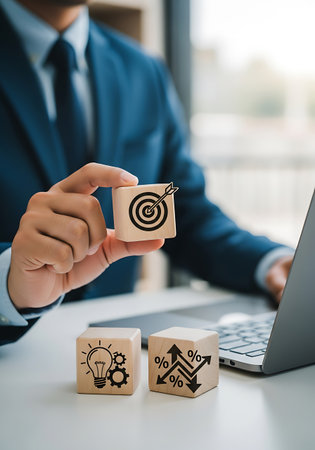 A businessman in a blue suit holds a wooden cube with a target and arrow symbol. Two other wooden cubes are on the desk, one with a lightbulb and gears, and the other with upward and downward arrows and percentage signs. A laptop is visible in the background.の素材
