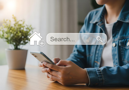 A woman uses a smartphone to search for a home. A search bar graphic with a house icon and magnifying glass is overlaid on the image.の素材