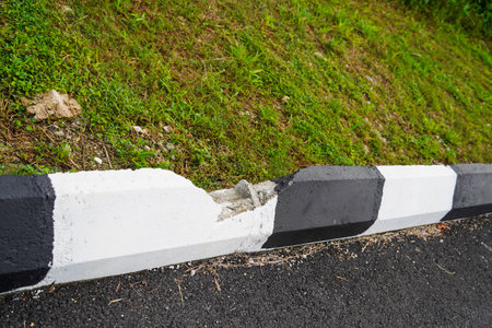Damaged black and white painted curb separating asphalt road from a grassy embankment.の写真素材