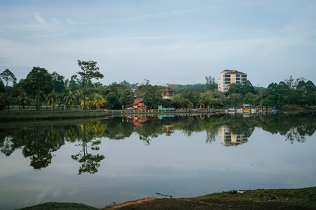 Serene lake reflection with lush green trees and buildings under a soft blue sky, perfect for nature and urban park concepts.の写真素材