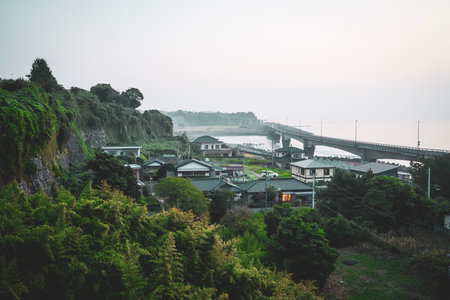 Coastal Japanese village with houses nestled against a green hillside next to a long bridge over the sea at dusk.の写真素材
