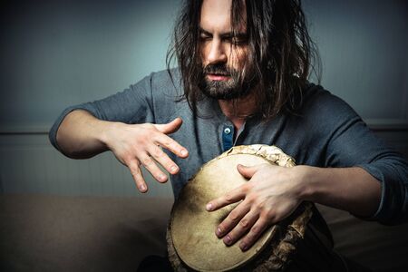 Long-haired man playing an ethnic percussion musical instrument jembe. Drummer playing african music