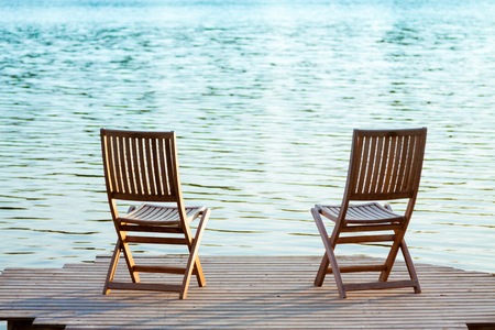 Two adirondack wooden chairs on dock facing a blue lakeの写真素材